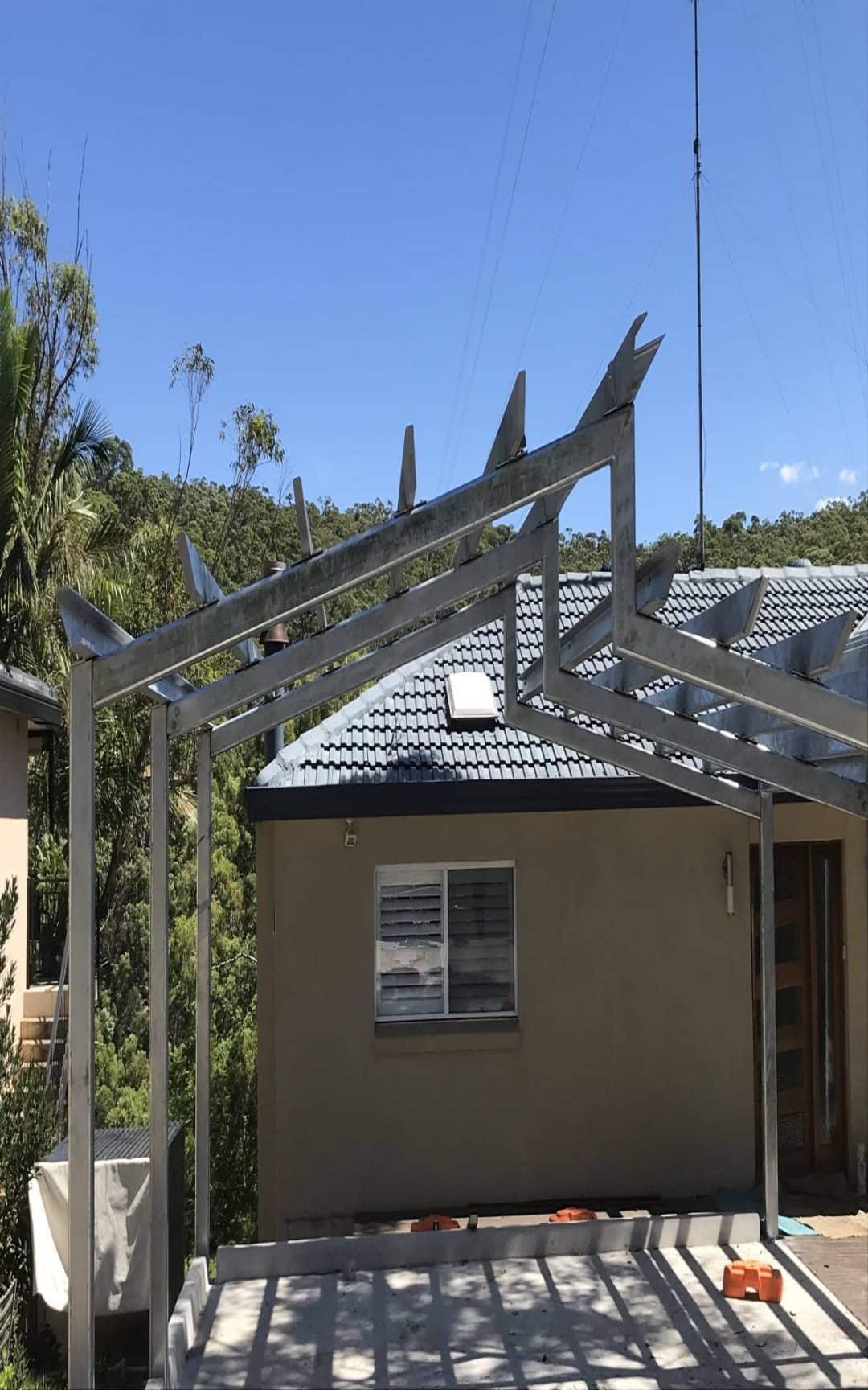 A steel frame for a new structure is being built over an existing patio next to a house with a tiled roof — Highlander Metals in Kirrawee, NSW