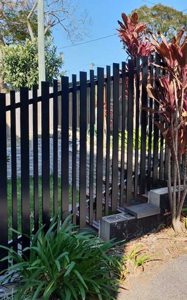 A black vertical slat fence sits atop a terraced stone wall, bordered by greenery and red-leafed plants — Highlander Metals in Kirrawee, NSW