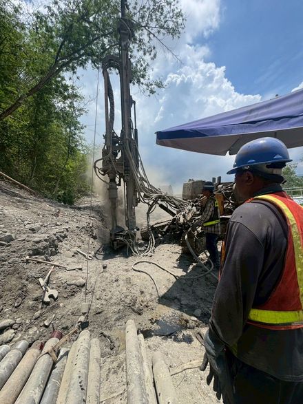 一名建築工人正在觀察鑽井平台。戶外環境塵土飛揚，天空湛藍。工人戴著安全帽，穿著安全背心。