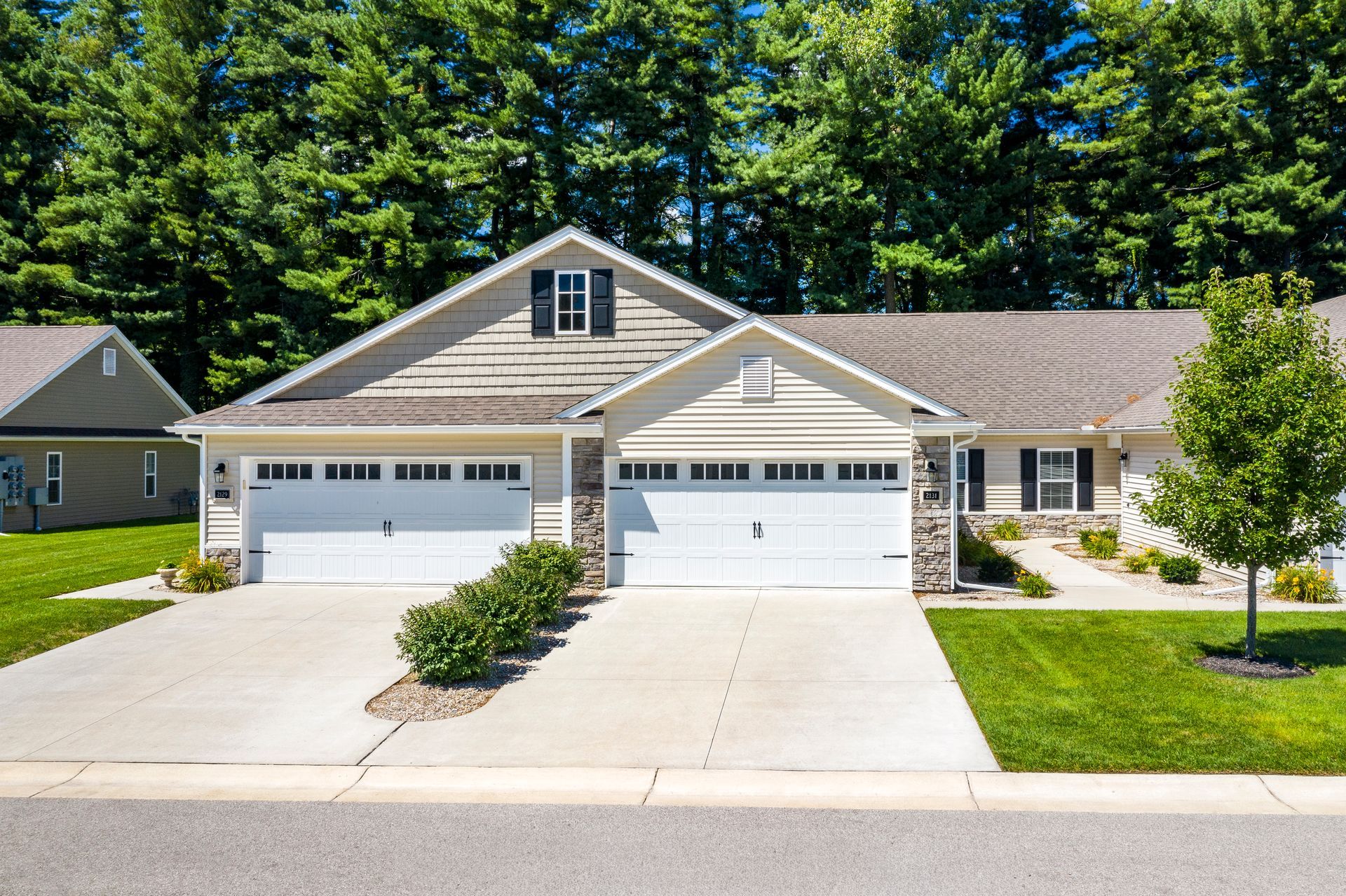 Tan and white suburban house with two garage doors and a concrete driveway, set against a backdrop of green trees.