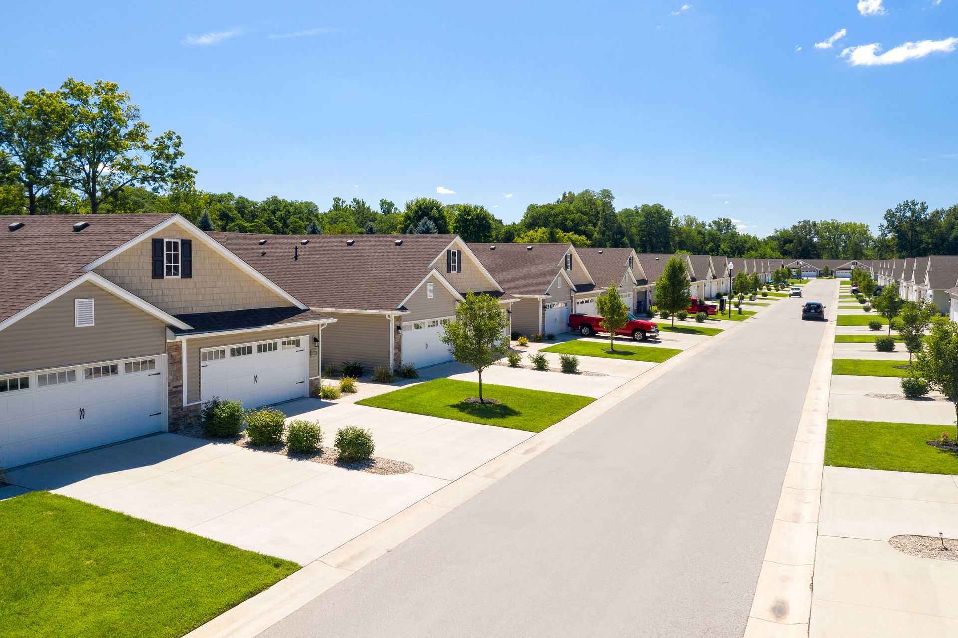 Row of beige houses with white garage doors, green lawns, and a paved road under a blue sky.