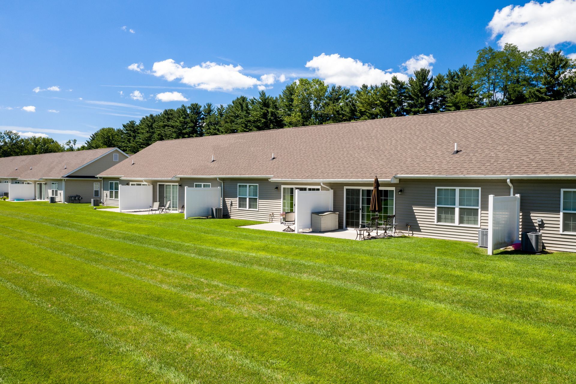 Row of single-story gray homes with patios and brown roofs, set in a grassy yard under a blue sky.