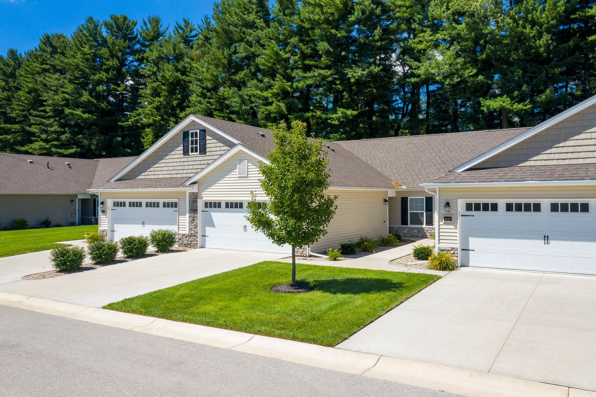 Row of beige townhouses with white garages, green lawn, and tall trees.