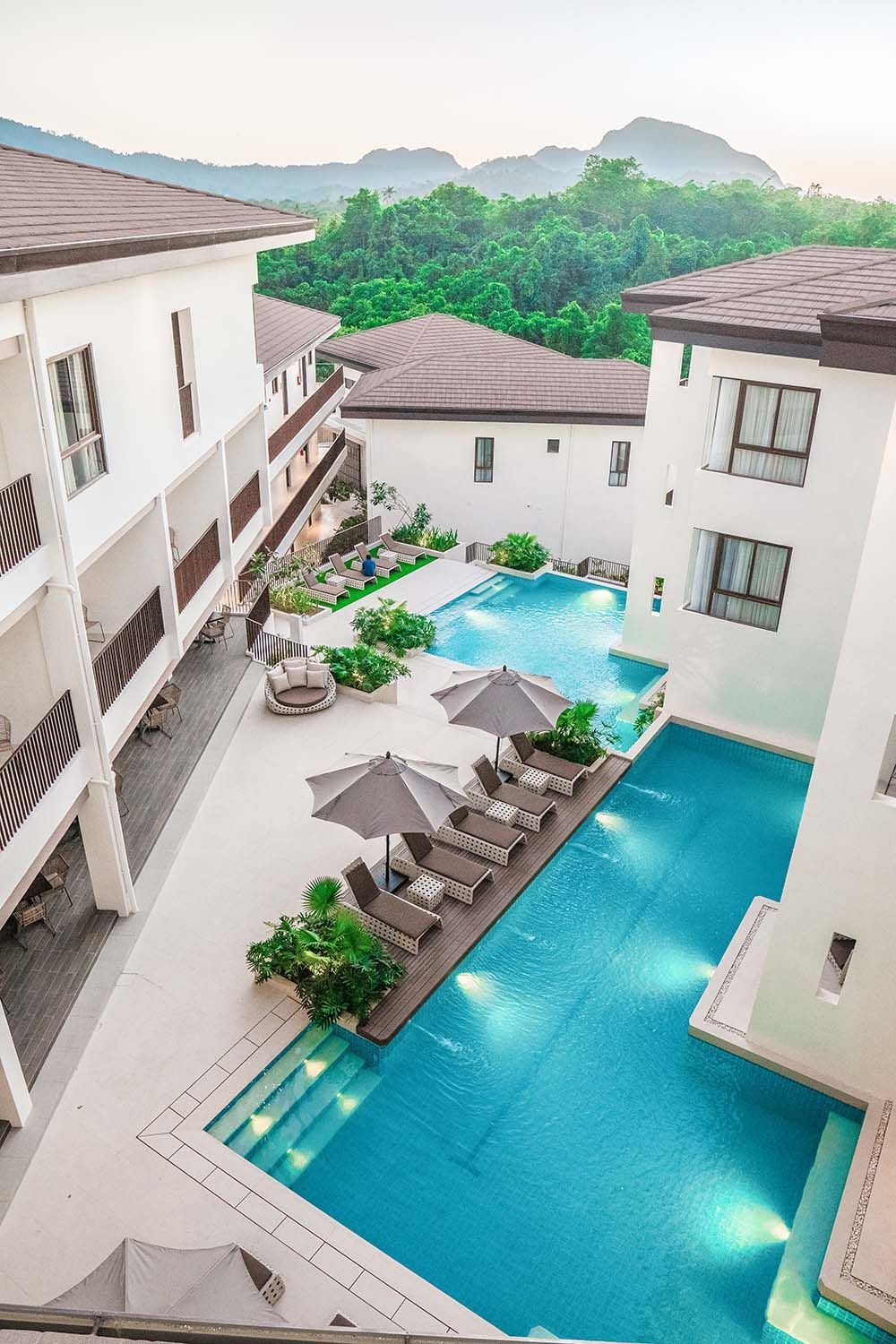 Courtyard with pool, lounge chairs, and buildings with brown roofs, trees in the background.