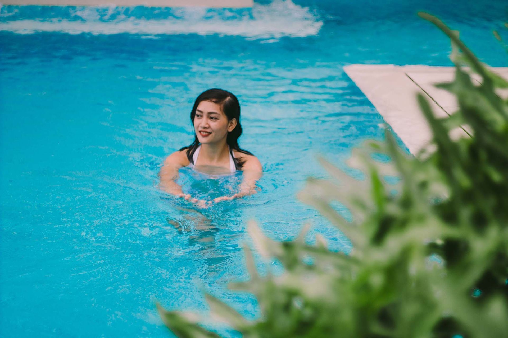 Woman in a white swimsuit smiles in a blue pool, with green foliage in the foreground.