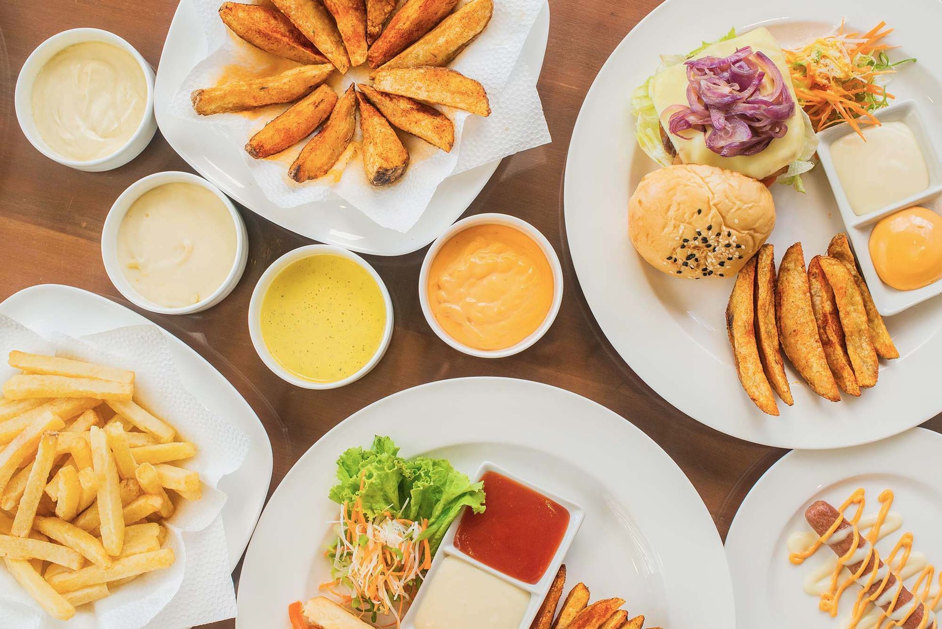 Plates of fries, potato wedges, burgers, and hotdogs with various dipping sauces on a table.