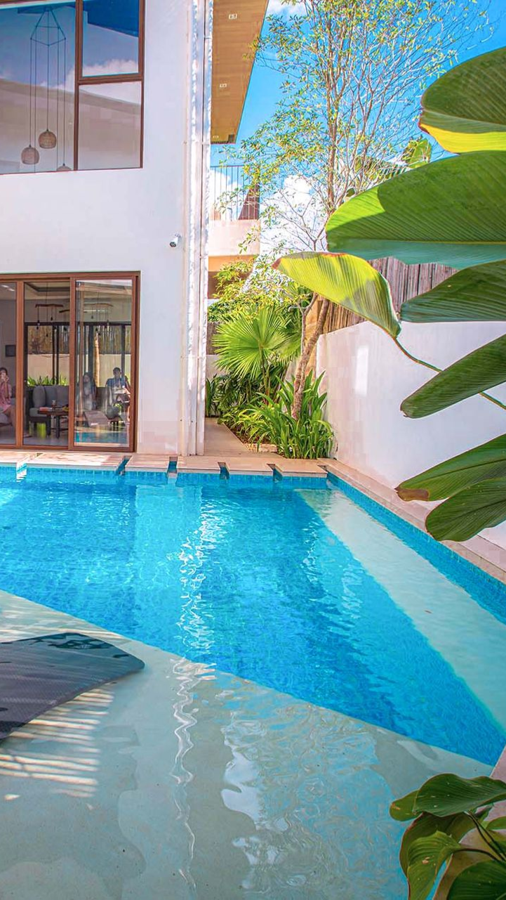 Swimming pool in courtyard next to white building with sliding glass door; lush greenery and blue sky.