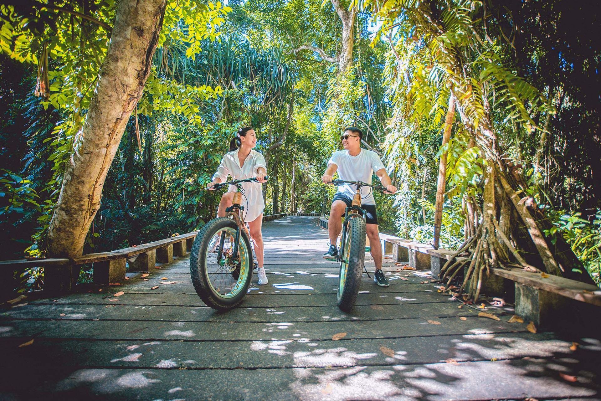 Two cyclists riding through a sunlit forest path with dappled shadows