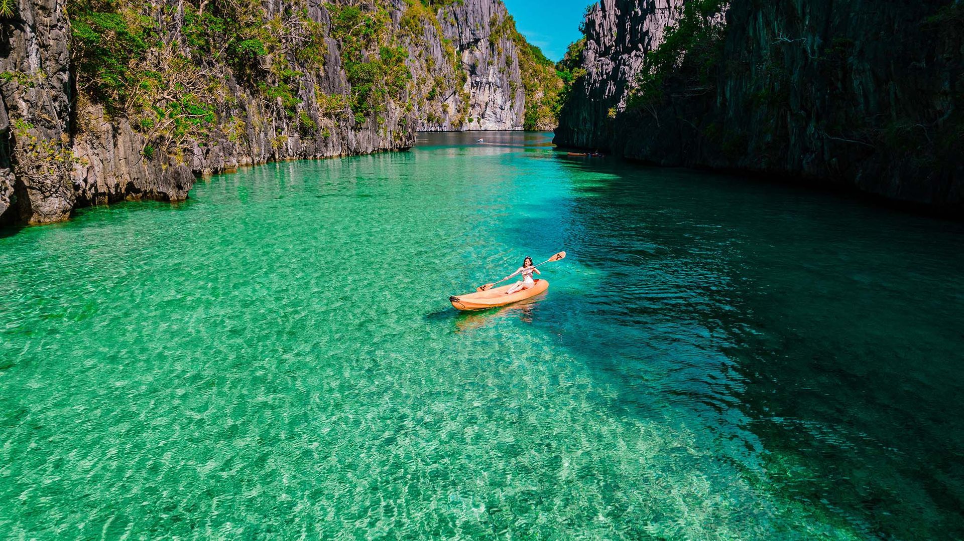 Kayakers on turquoise water between rocky cliffs. Lush green vegetation and blue sky.