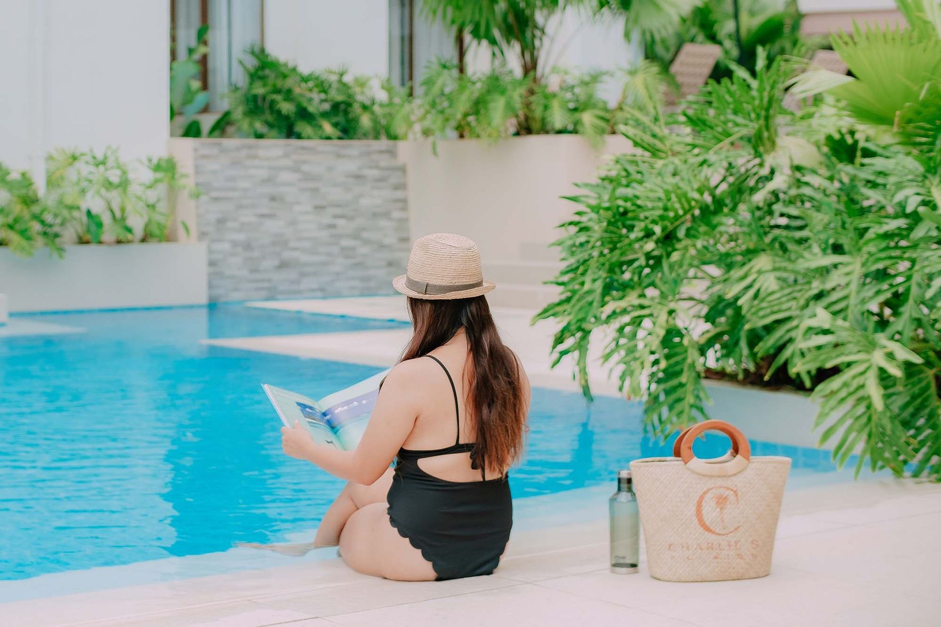 Woman in swimsuit and hat by pool, reading a book. A bag and water bottle sit nearby.