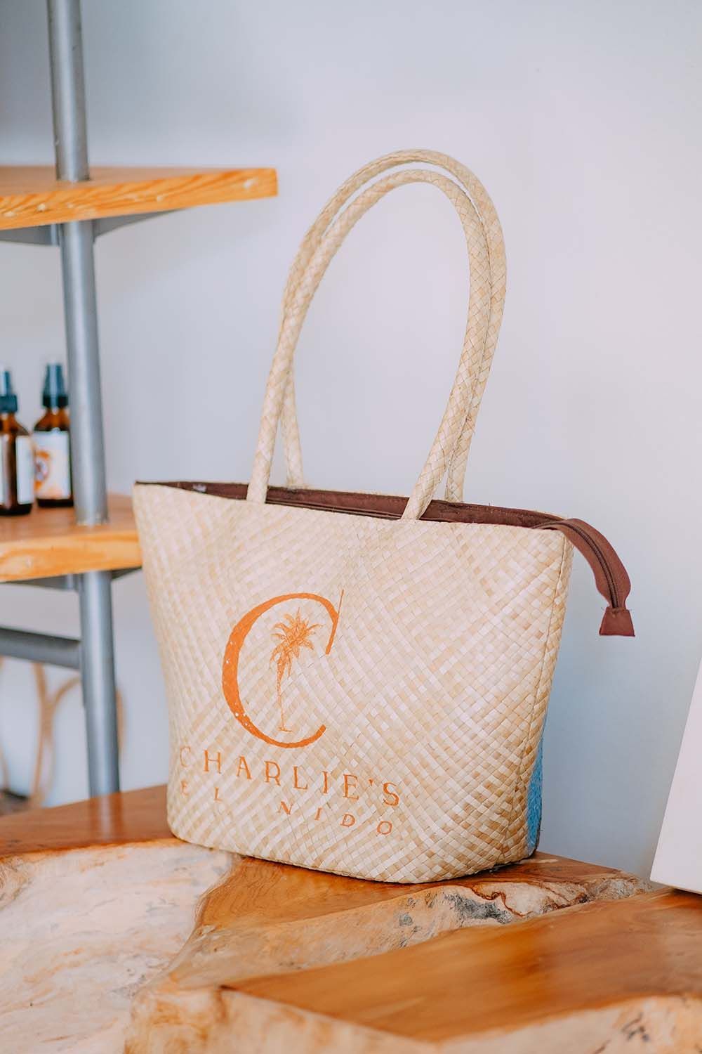 Woven tote bag with tan handles and logo, sitting on a wooden surface near shelves.
