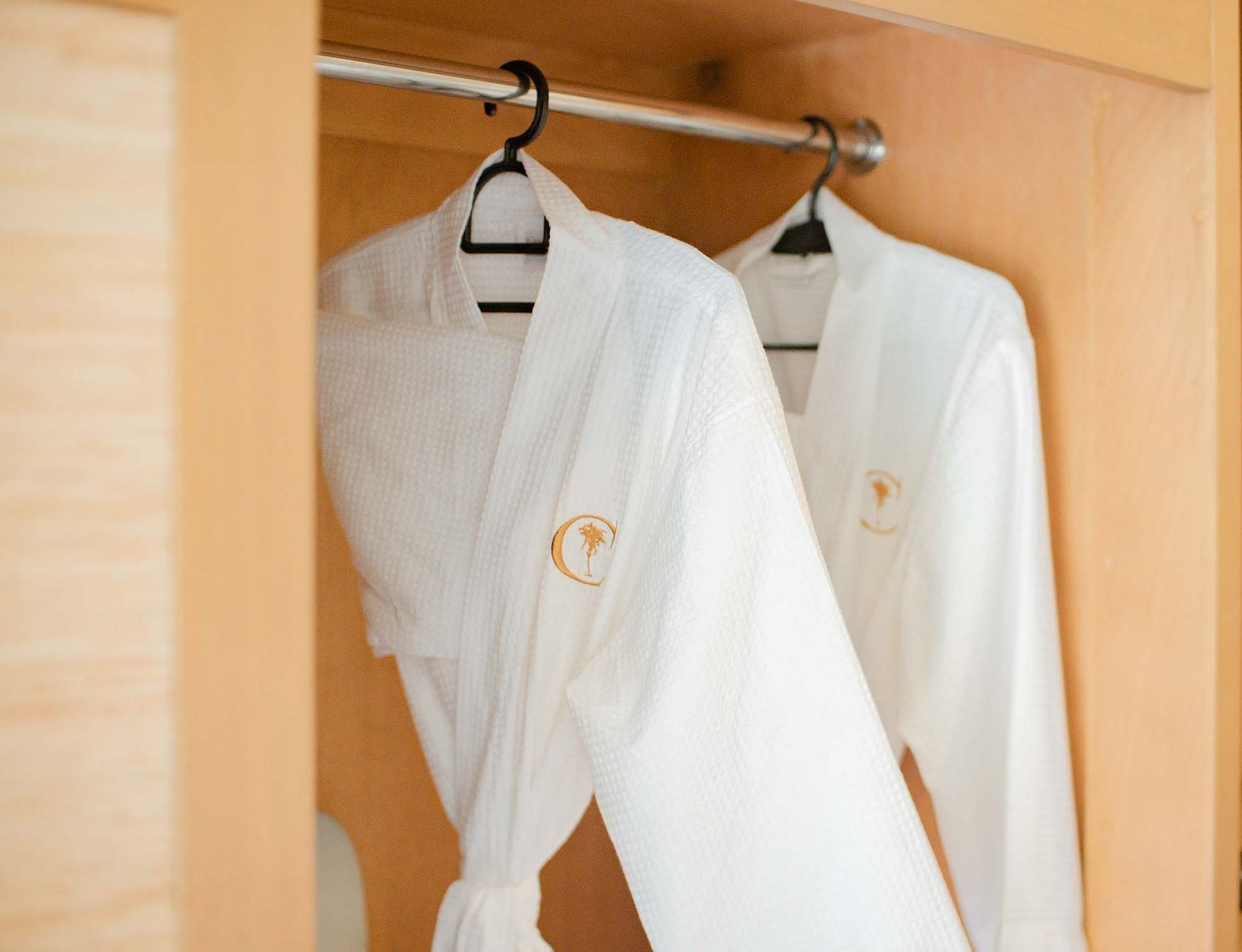Two plush white bathrobes with embroidered logos hanging in a wooden wardrobe
