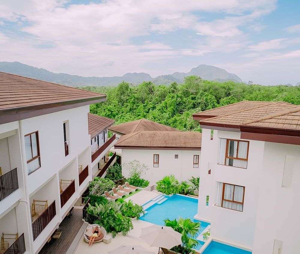Resort buildings with pool, lush greenery, and mountain backdrop under a cloudy sky.