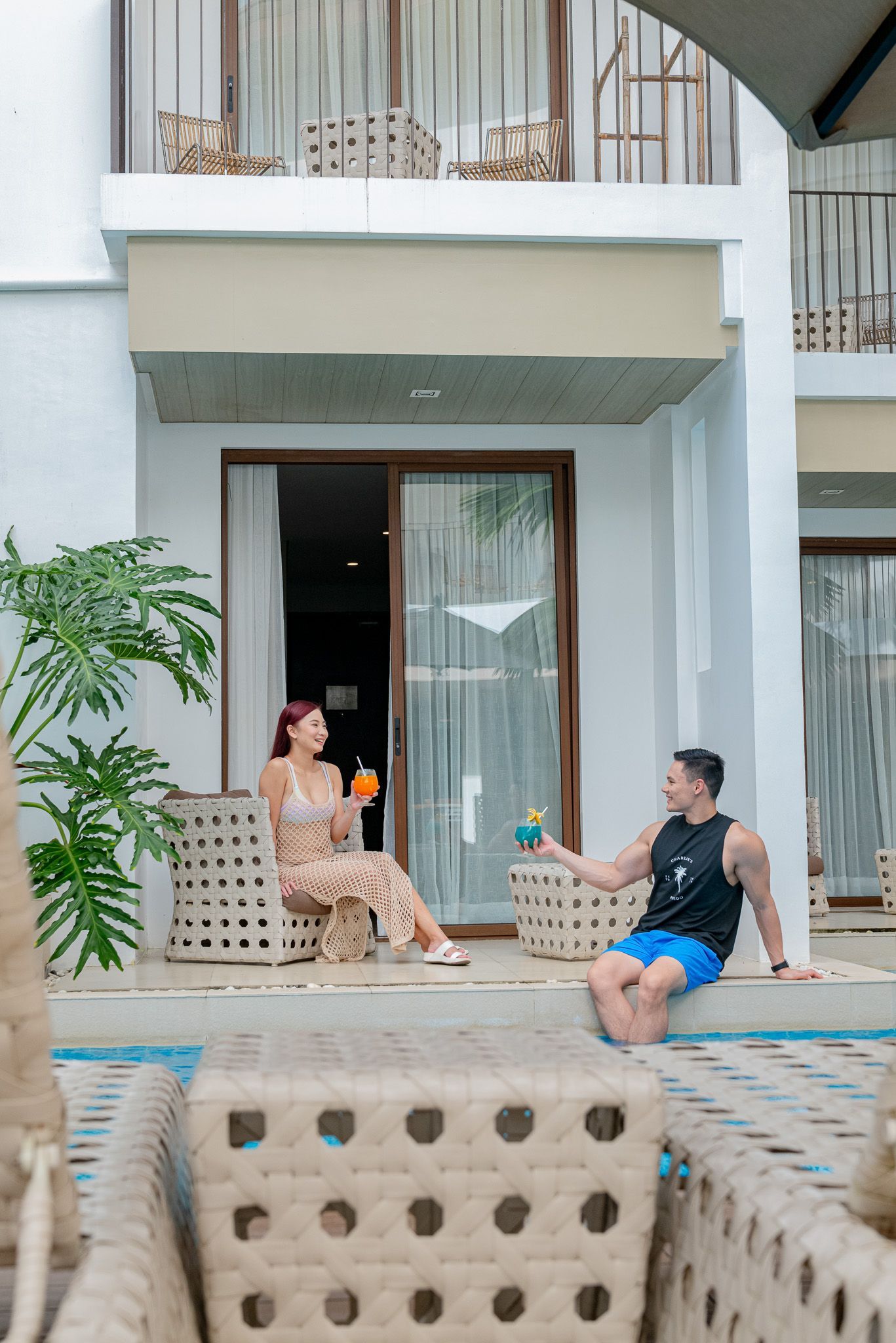 Couple toasting drinks on a patio. White building, glass door, rattan furniture, greenery.