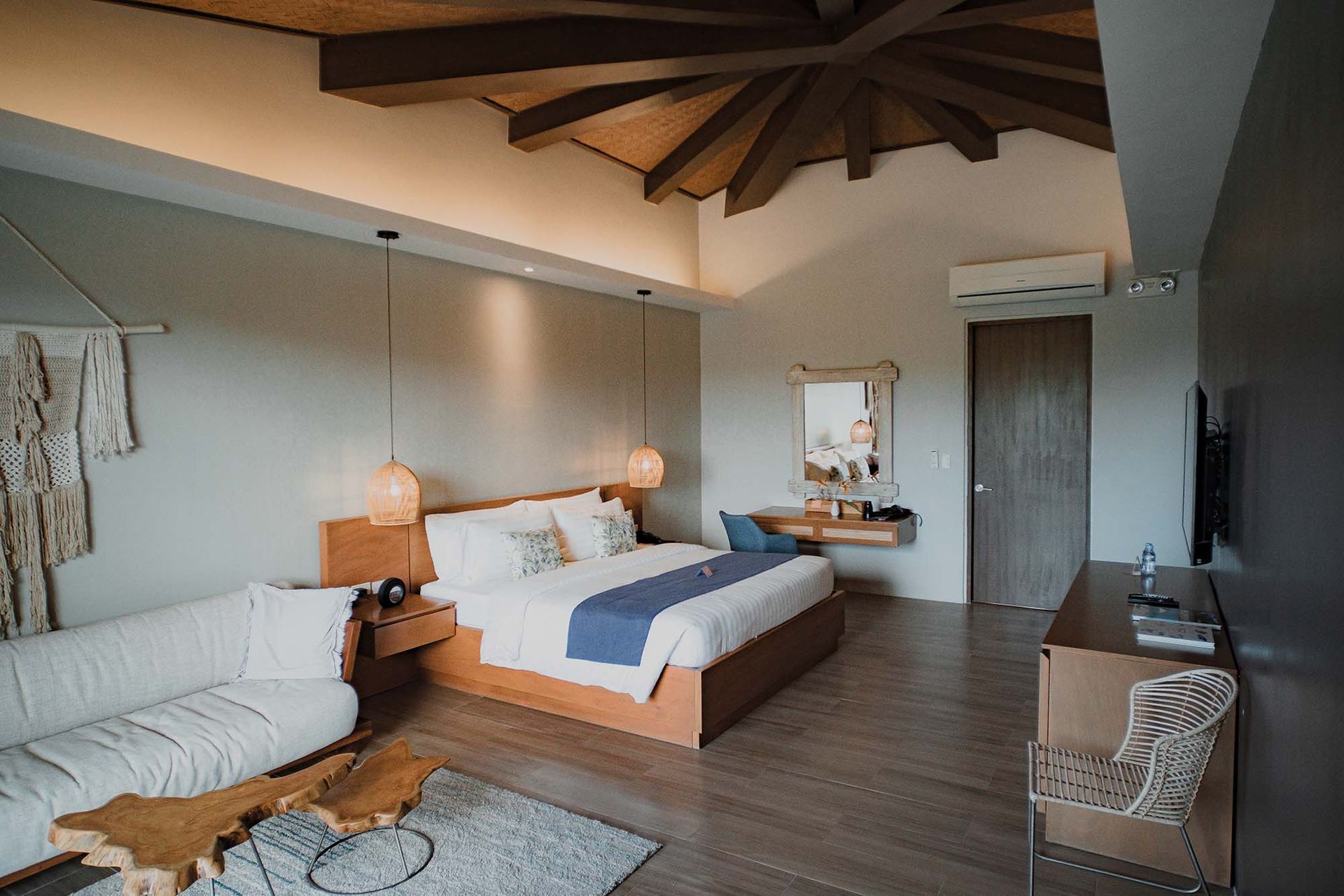Spacious hotel room with wooden beams, king bed, sofa, desk, and TV. Neutral colors, natural light.