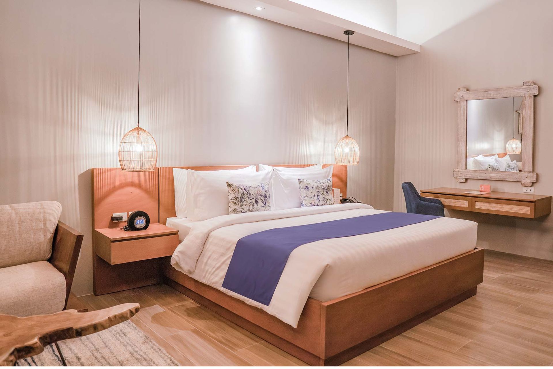 Bedroom with king bed, floating nightstand, small chair, desk with mirror; light wood and neutral colors.