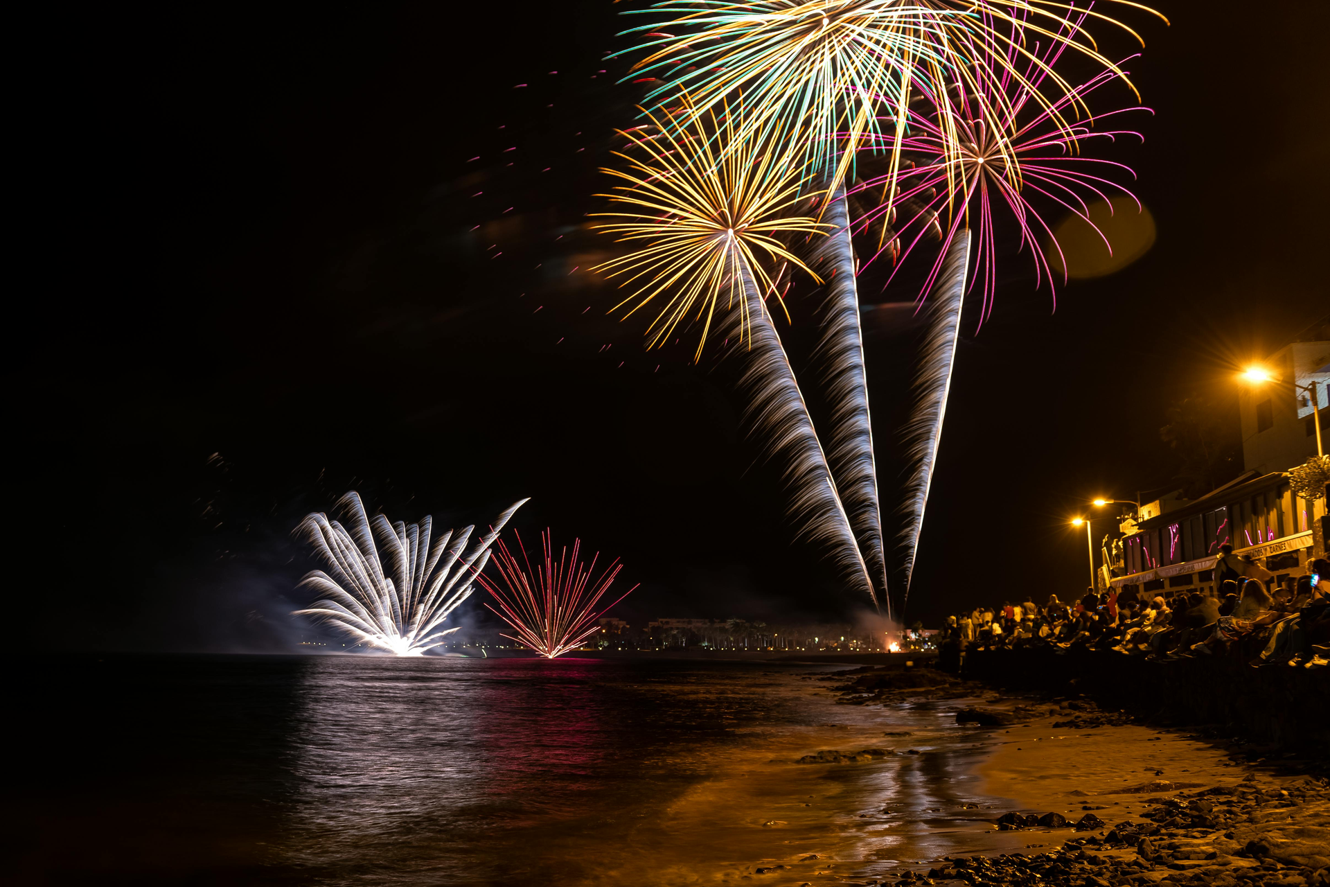 Fireworks erupting over the ocean at night, with spectators on the shore.