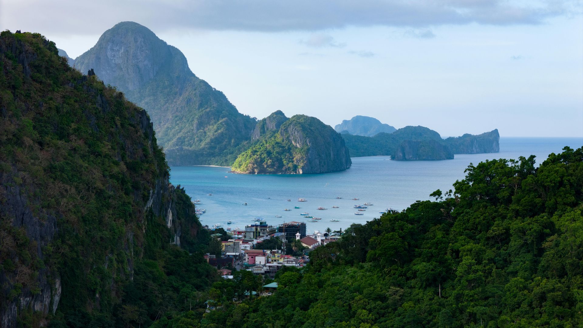 Bay with mountains, boats, and town, framed by green trees and cliffs under a blue sky.