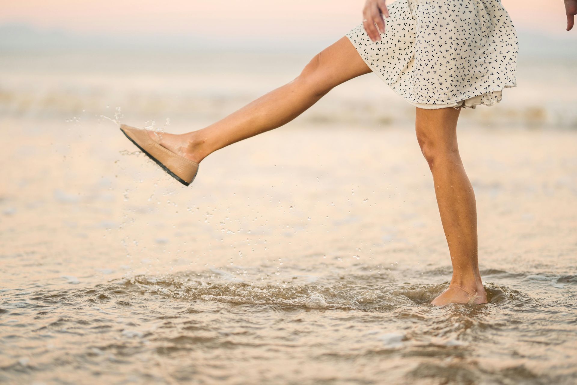 Woman kicks foot in ocean water, splashing. She wears a polka dot dress and a shoe.