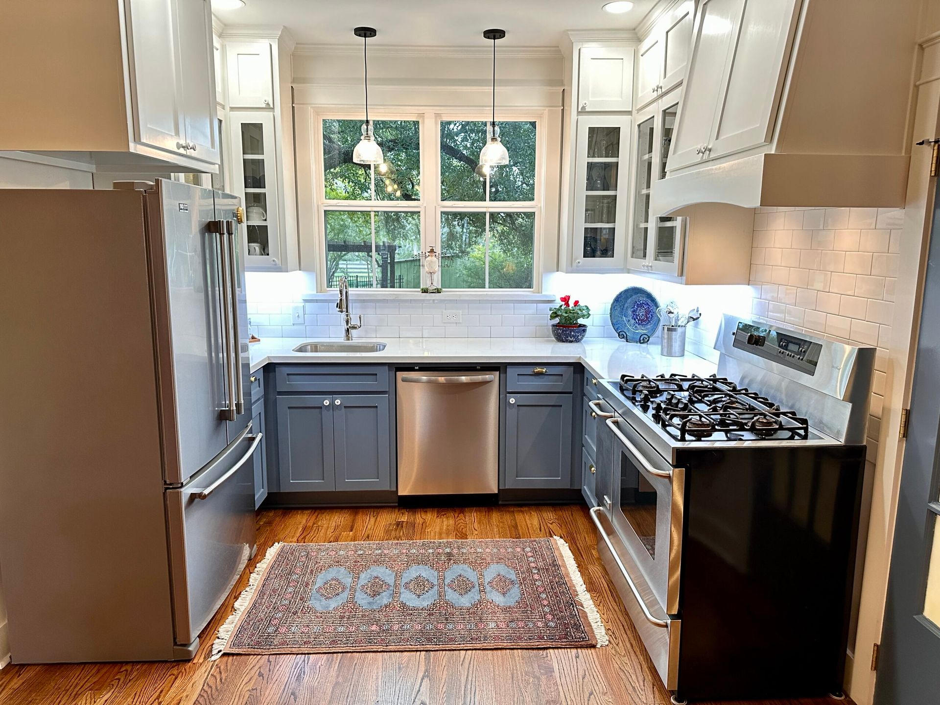 A kitchen with blue cabinets , stainless steel appliances , a rug and a large window.