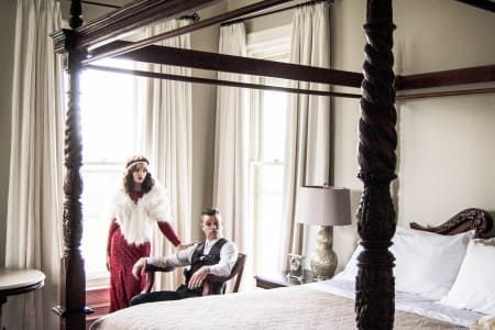 A man and a woman are standing next to a four poster bed in a bedroom.