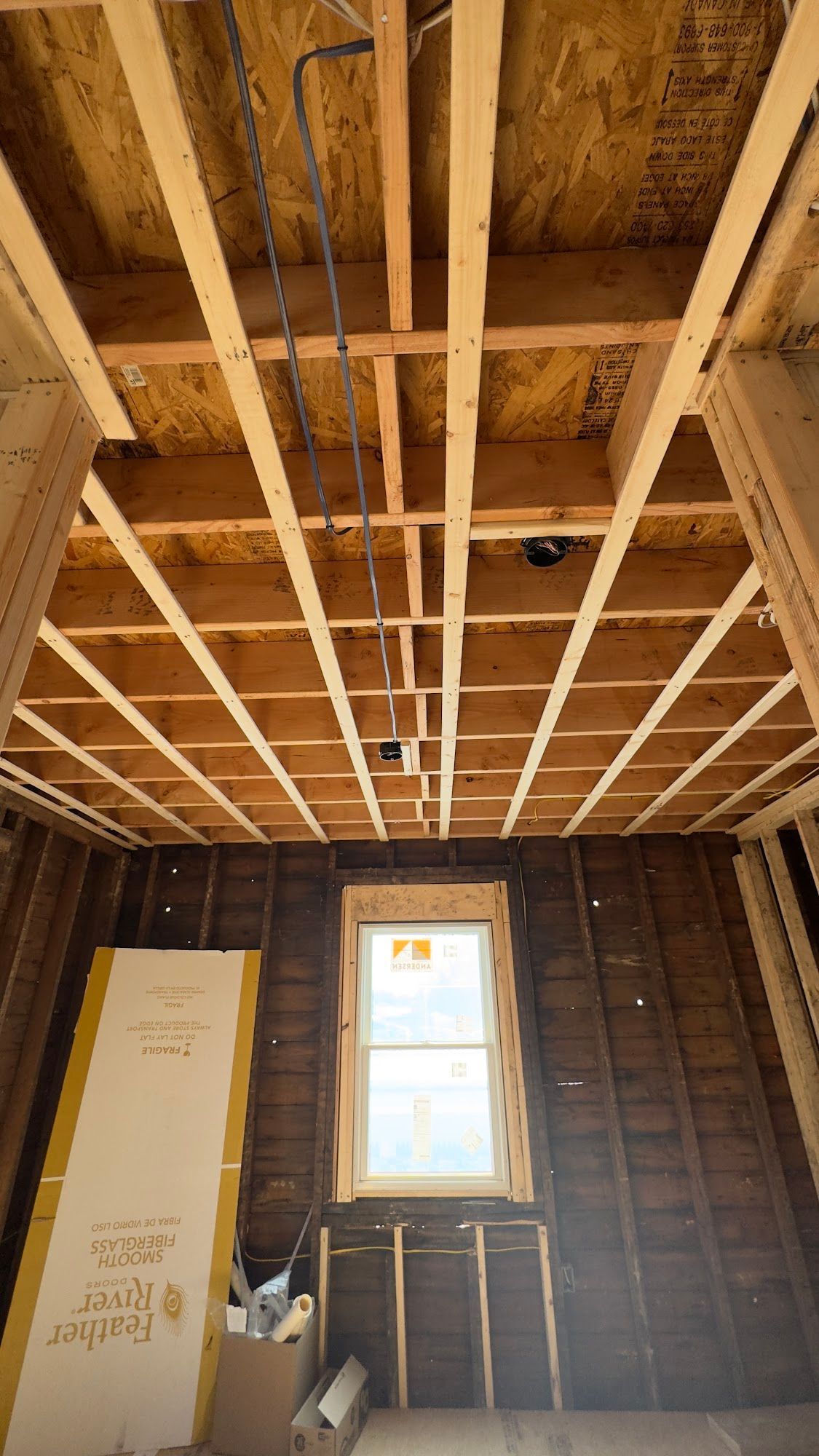 Interior of a room under construction, showing exposed wood beams and wiring in the ceiling, a window.