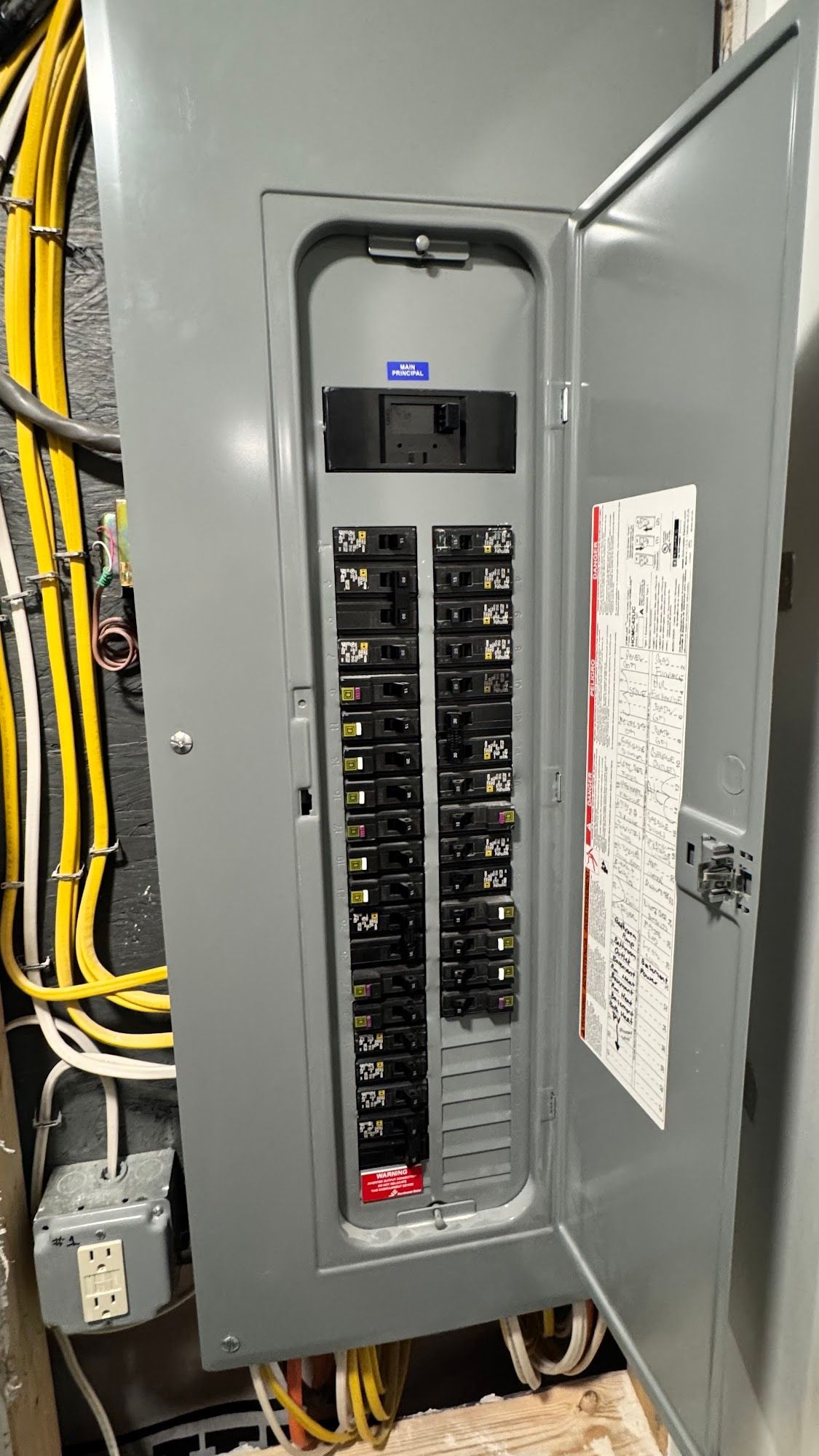 Open gray electrical panel with various circuit breakers. Wires and a small electrical box are visible nearby.
