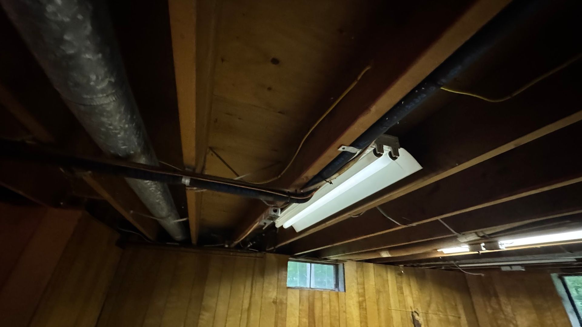 Basement ceiling with wood paneling, pipes, and a fluorescent light fixture.
