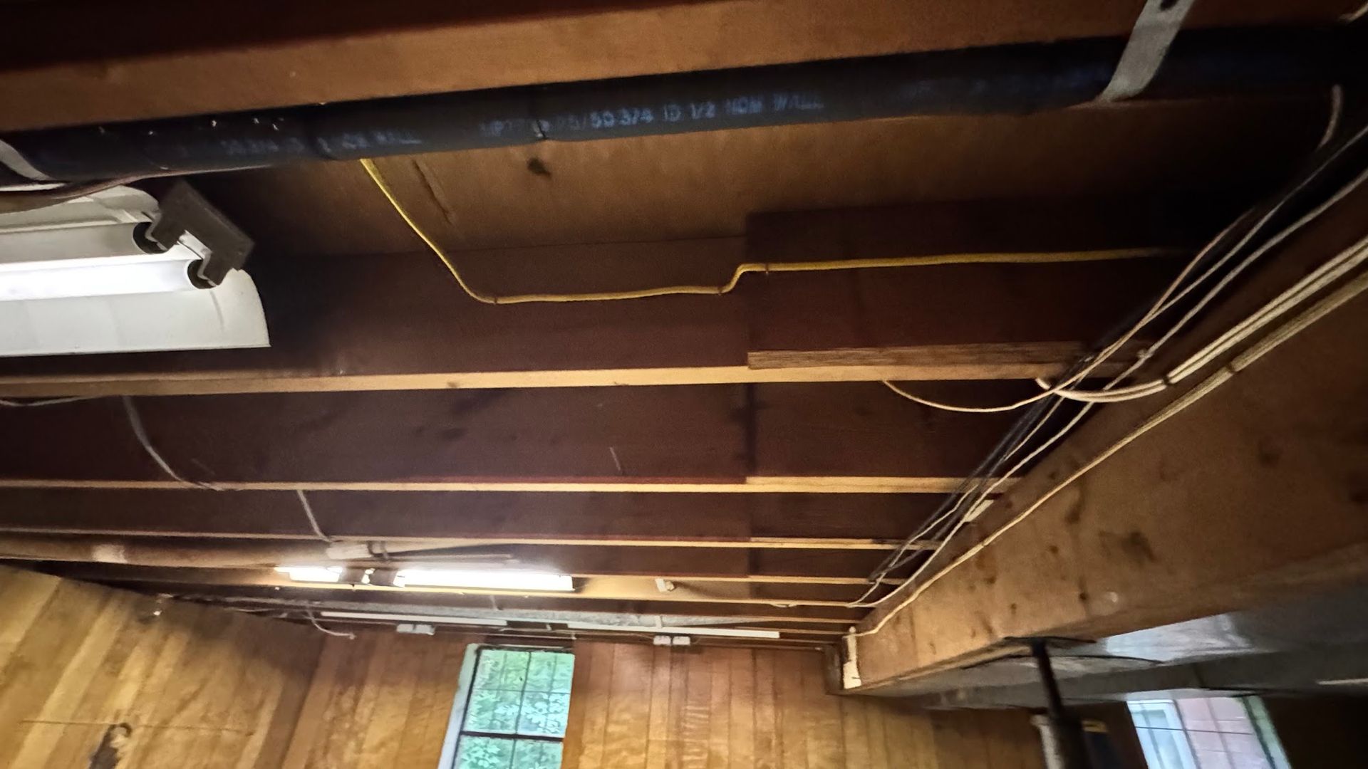 Basement ceiling with exposed beams, pipes, and wiring. Wood paneling on walls, fluorescent light fixture.