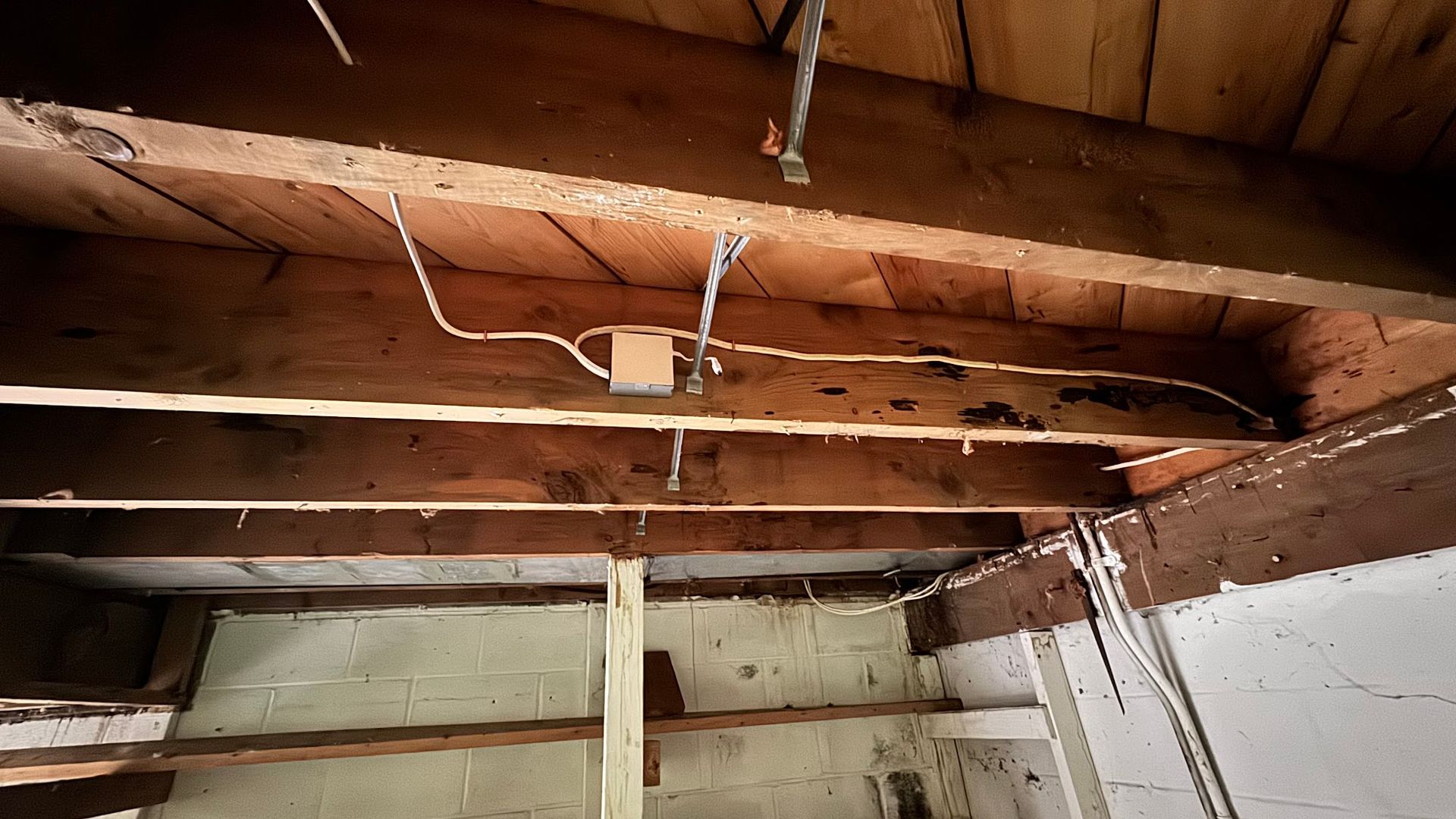 Wooden beams with exposed wiring and electrical box in a dimly lit, unfinished interior.