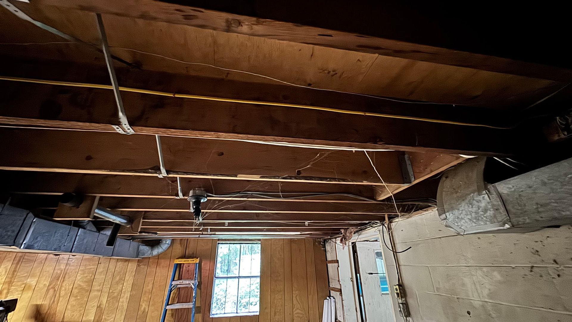 Basement ceiling with exposed beams, electrical wiring, ductwork, and window. A ladder stands nearby.