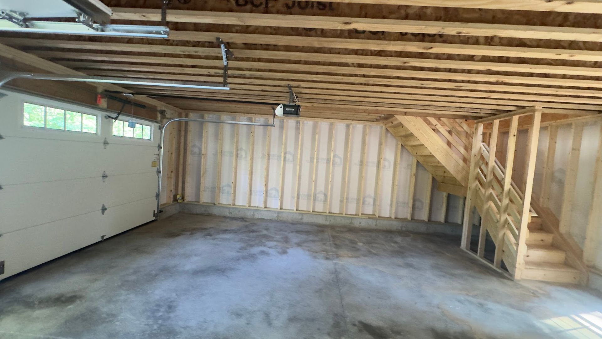 Unfinished garage with concrete floor, wooden walls, and staircase; garage door on the left.