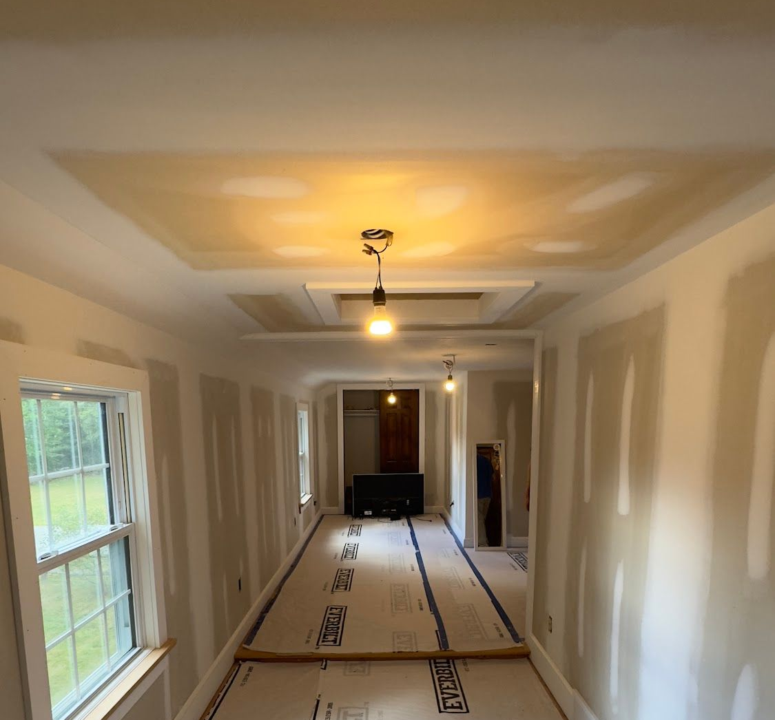 Electrical box and conduit on the side of a beige house with a window above. Green plants grow along the foundation.