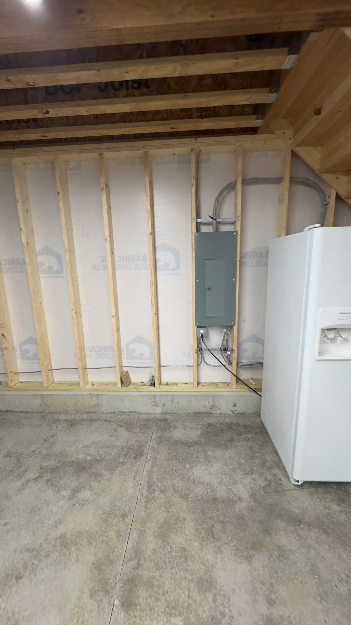Gray electrical panel on a wood-framed wall in a garage, next to a white refrigerator.