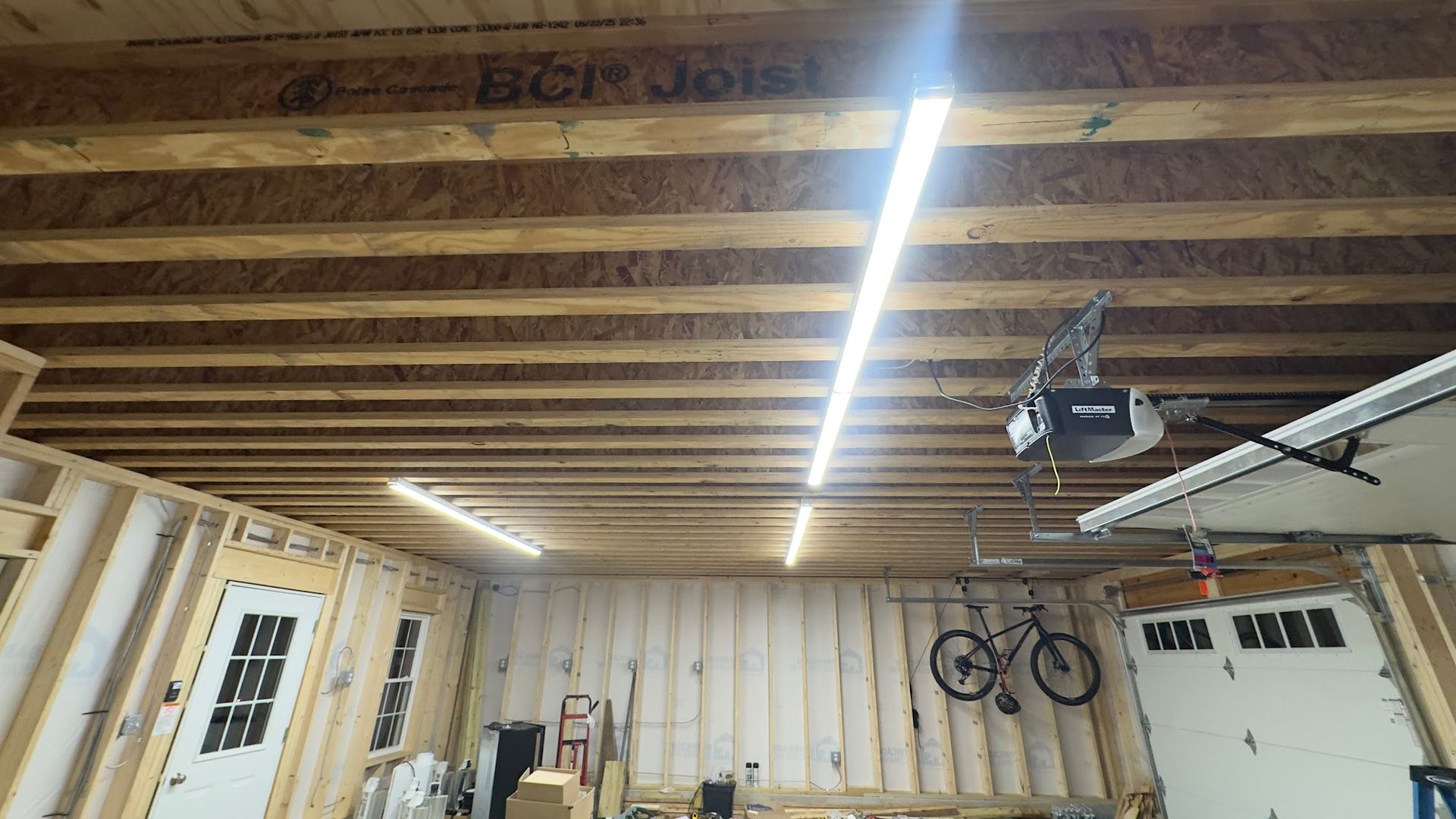 Garage interior with wooden ceiling beams, strip lights, and a closed garage door. A bicycle hangs from the ceiling.