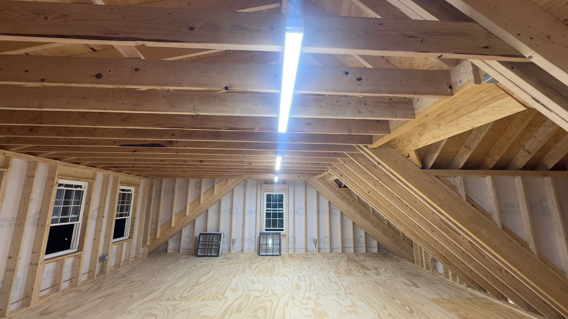 Interior view of an unfinished attic space. Exposed wooden framing with windows and a single fluorescent light fixture.
