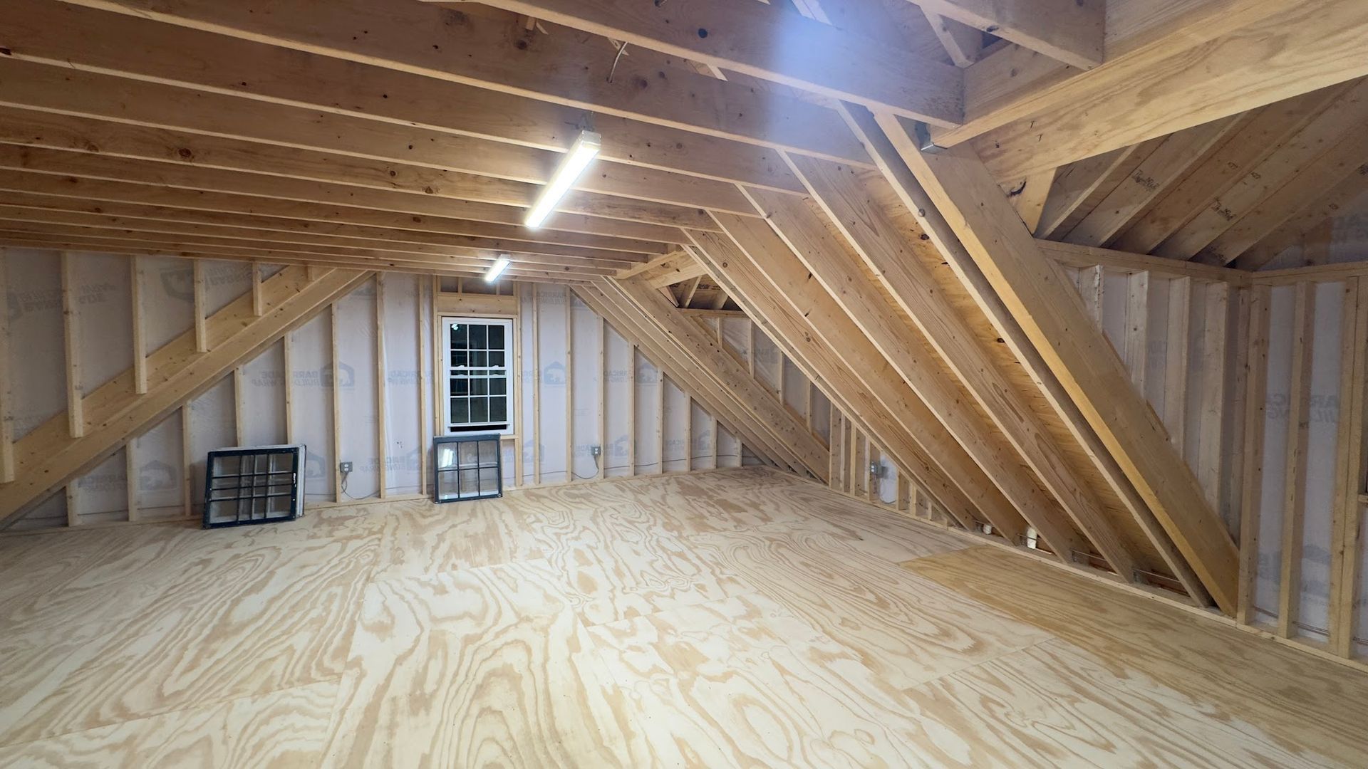 Interior view of an unfinished attic with wooden framing, plywood floor, and a small window.