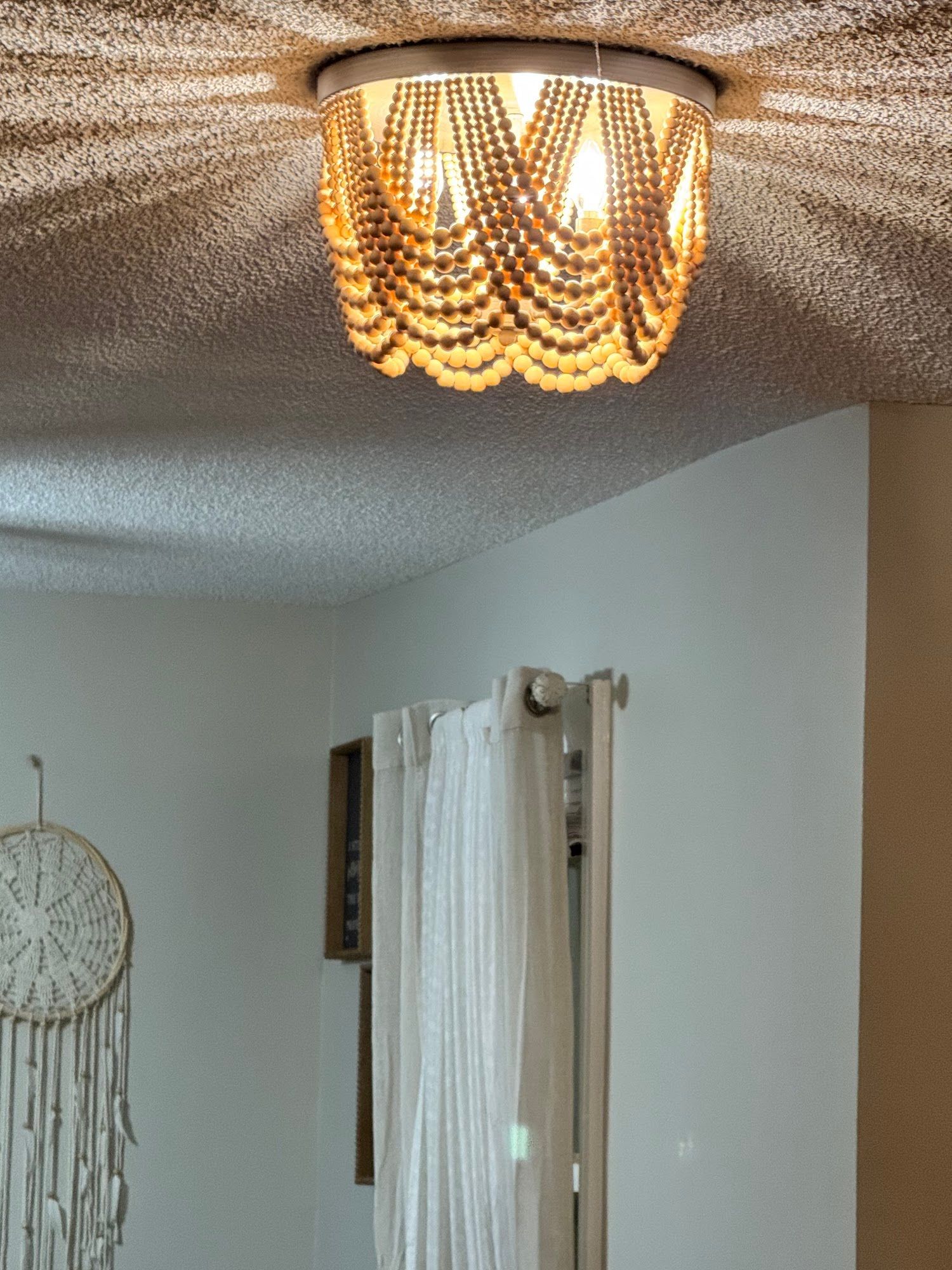 Ceiling light with wooden bead shade, illuminating a room with white walls and sheer curtains.