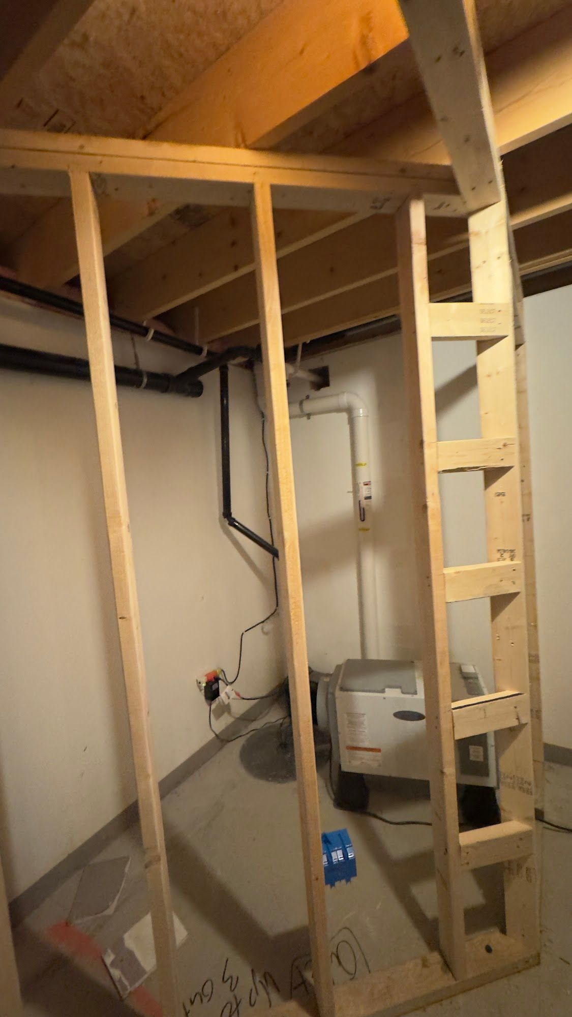 Framing of a wall in a basement, showing pipes, wiring, and a water heater unit.