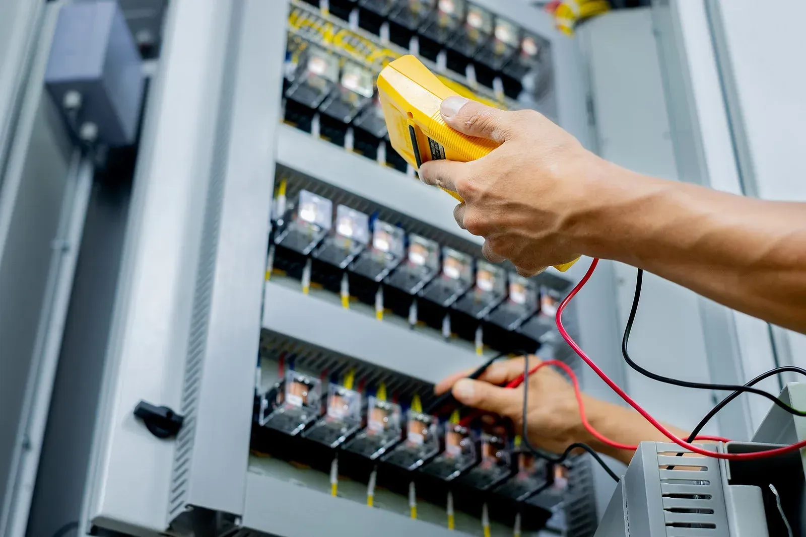 Person using a yellow multimeter to test electrical components inside a gray control panel.