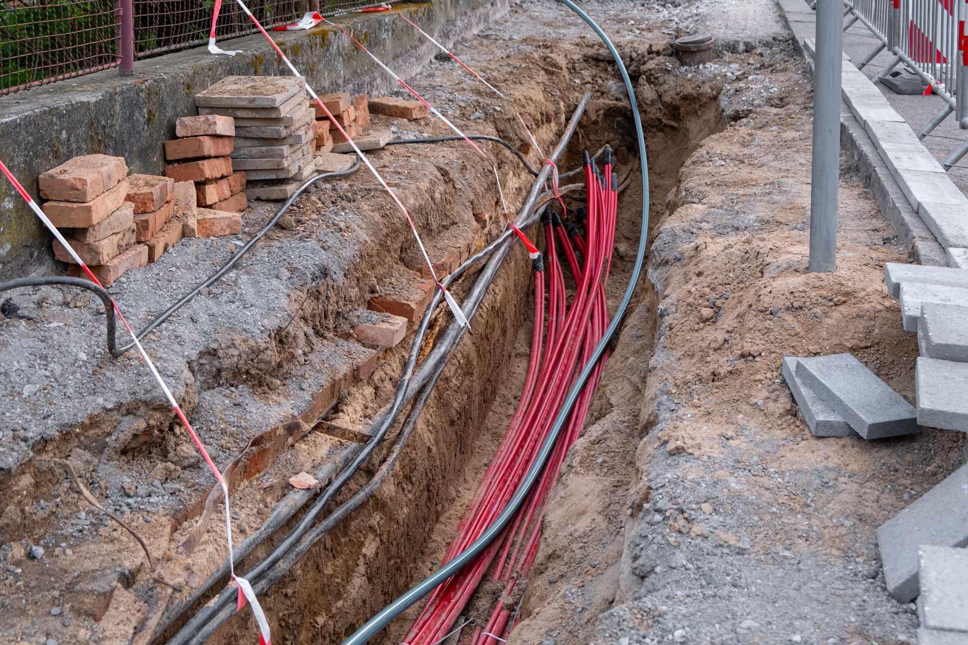 Roadside trench with exposed red cables and stacked bricks beside a construction area
