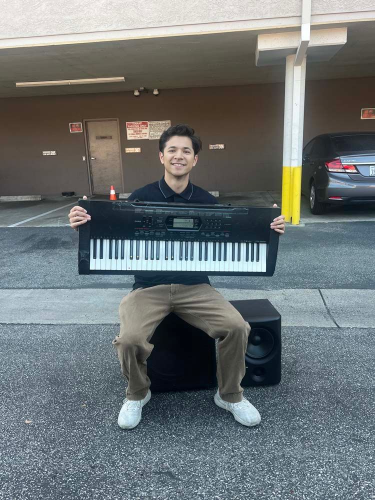 Person smiling and holding a keyboard in a parking garage, seated on a speaker box.