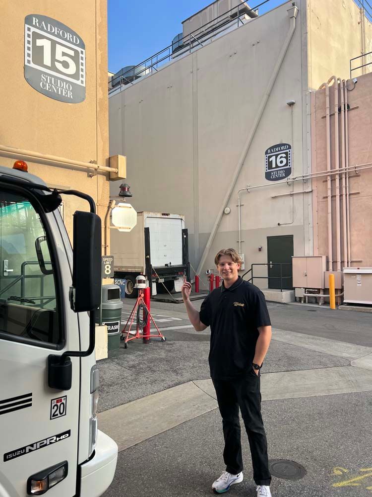 Person standing outside a large building, waving near a truck and a sign marked “16.”