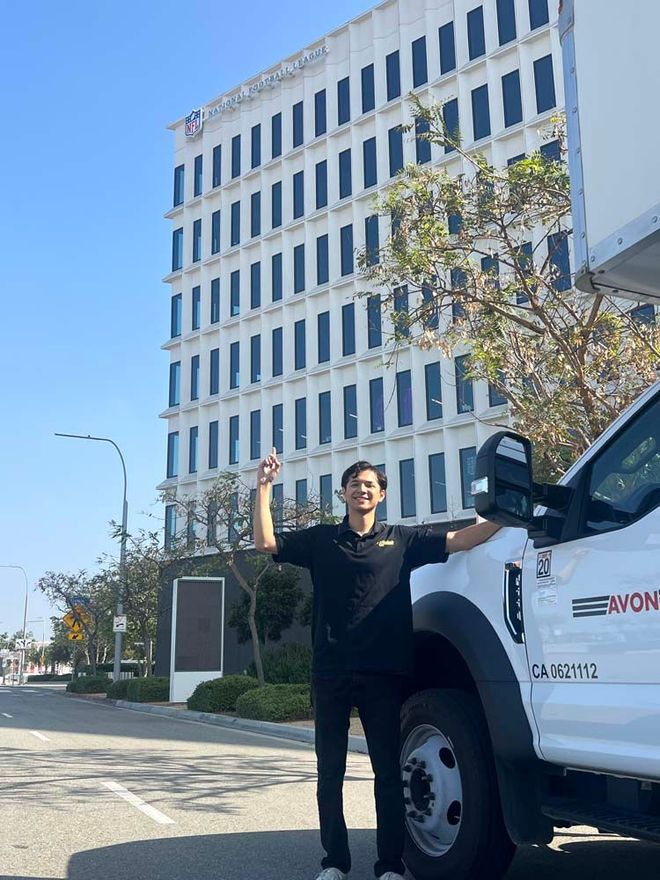 Person standing beside a white utility truck in front of a tall office building, waving under a clear blue sky.