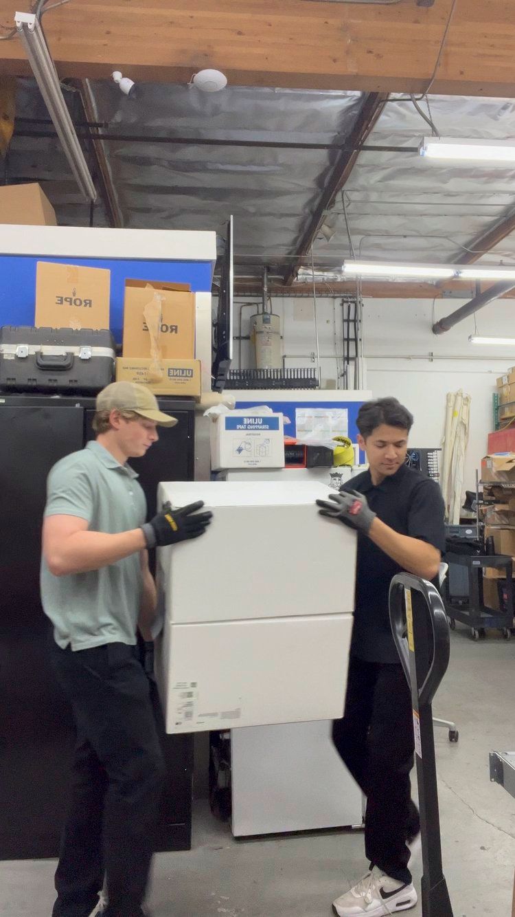 Two men are carrying a refrigerator in a warehouse.
