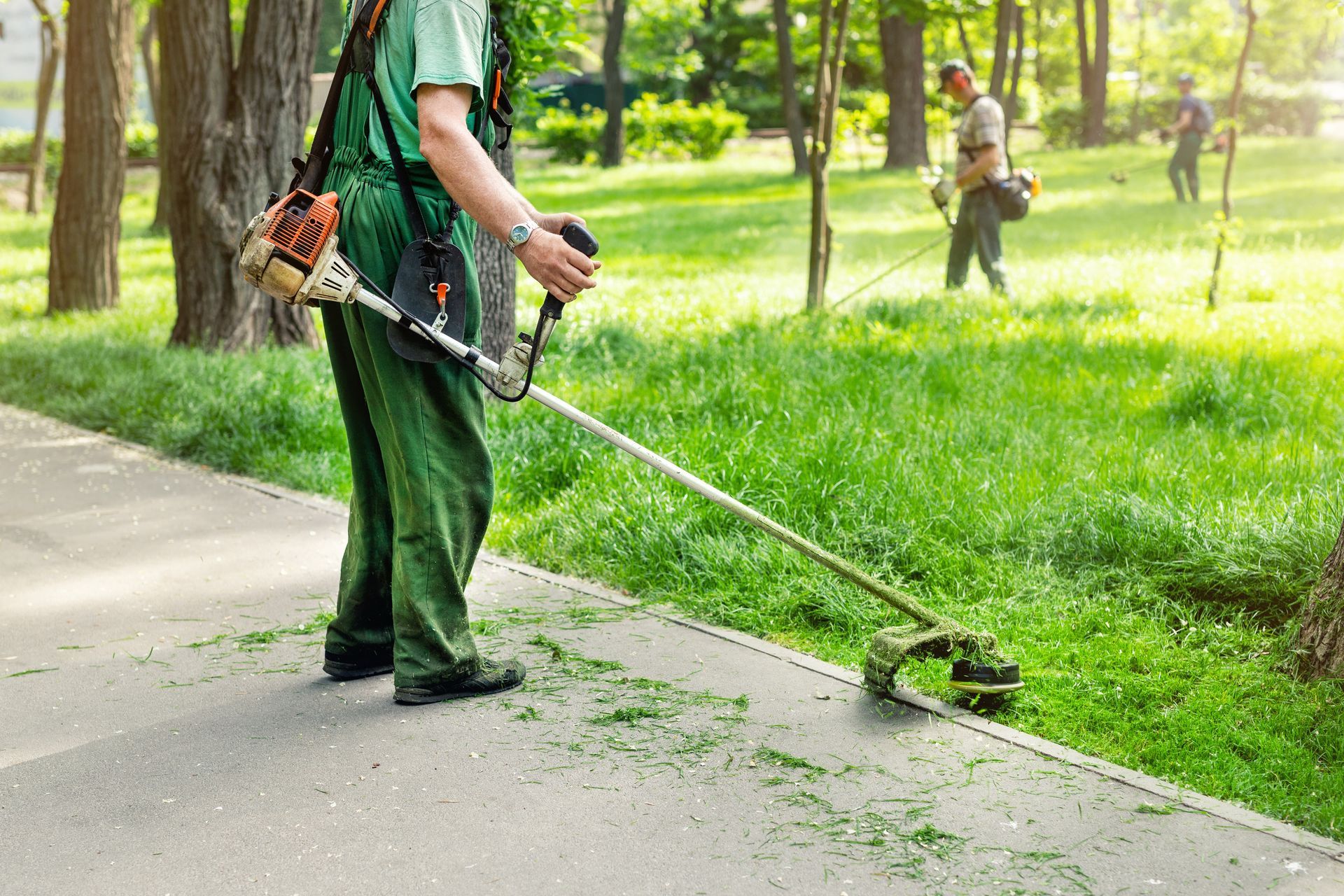 Man in green overalls using a weed wacker to trim grass along a sidewalk in a park.