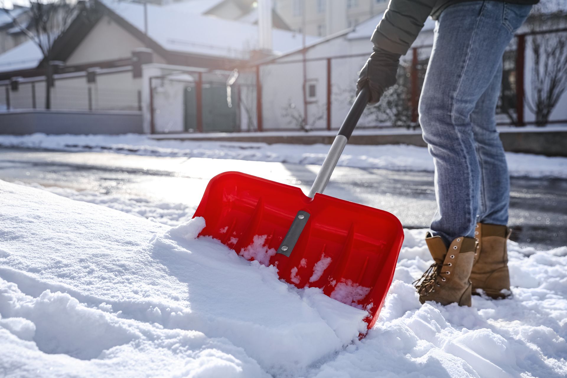 Person shoveling snow from a sidewalk with a red shovel on a sunny winter day.