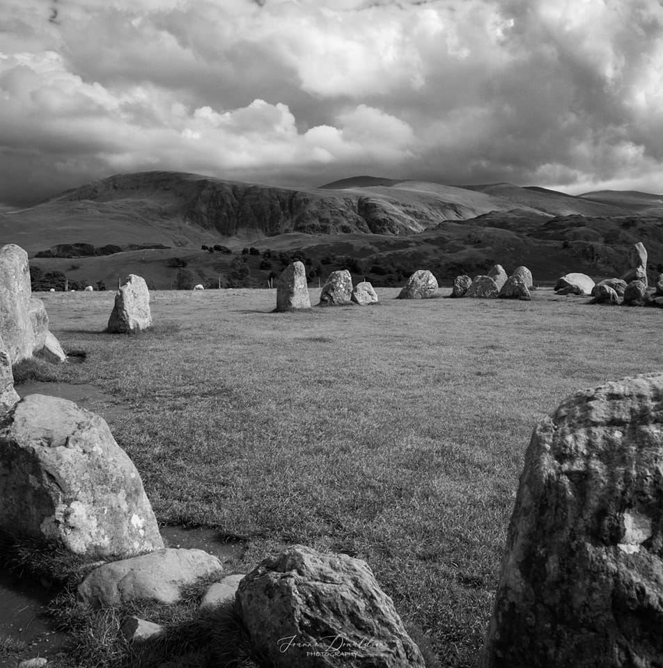 Stone circle in a grassy field with hills in the background under a cloudy sky.