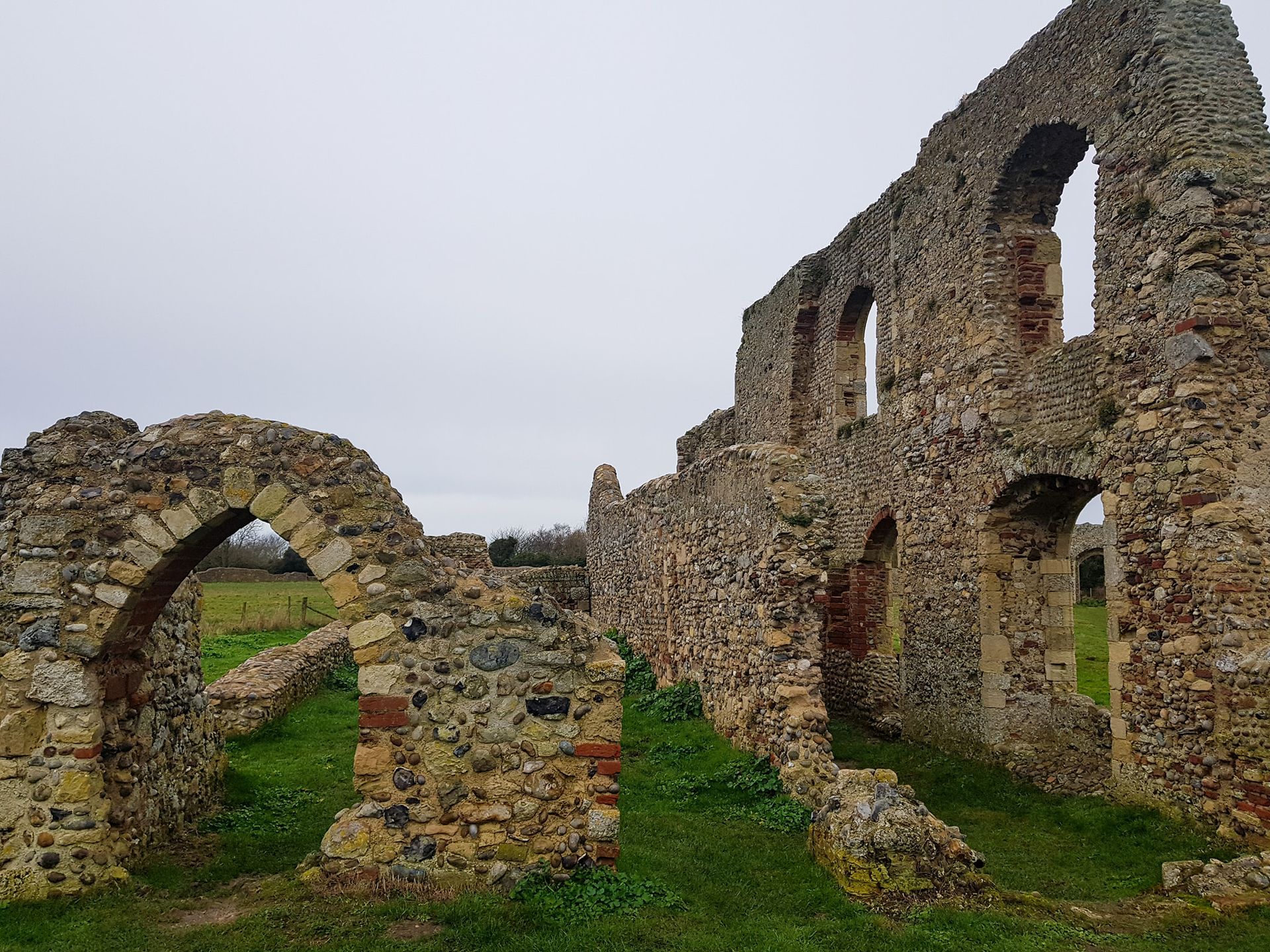 Ruins of a stone building with arched windows in a grassy field under a gray sky.