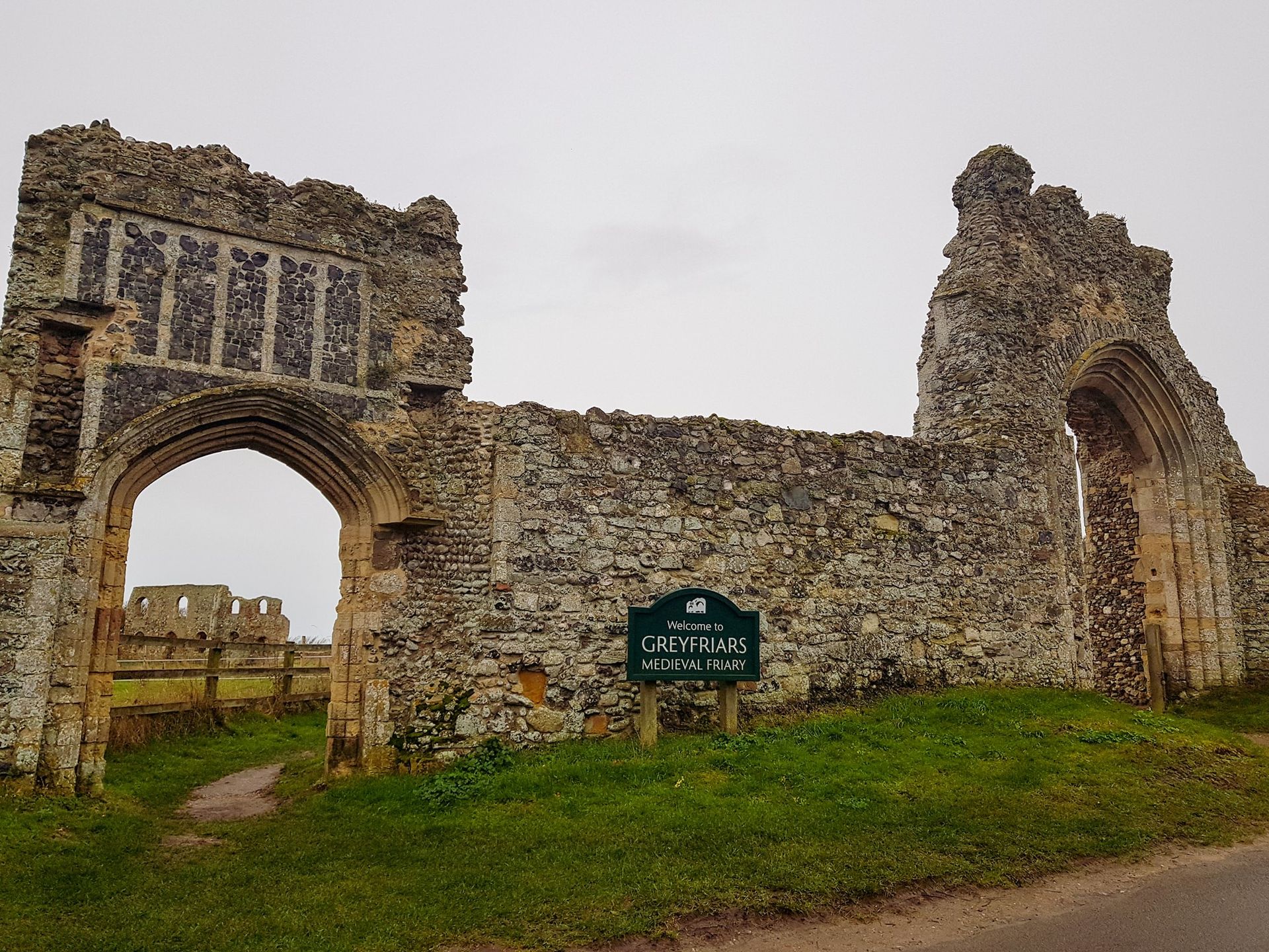 Ruined stone gate with sign, field and overcast sky.